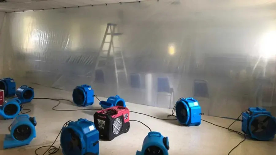 Blue air movers and a red dehumidifier drying an emptied room inside plastic containment walls